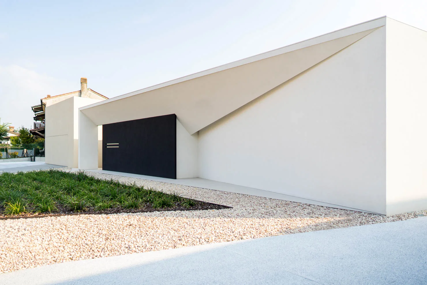 Architettura minimalista di Villa MD Home a Verona, caratterizzata da un design moderno con porta a filo muro nera, pareti bianche, tetto con elementi triangolari, ghiaia chiara e vegetazione ornamentale. Progetto esclusivo di Federico Cappellina & Partners Architects