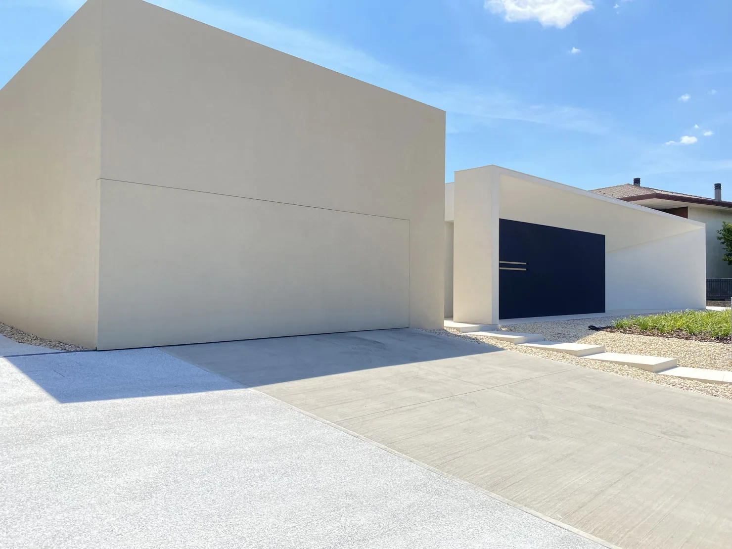 Architettura minimalista di Villa MD Home, Verona, con porta nera a filo muro, pareti bianche, tetto triangolare a sbalzo, ghiaia chiara e vegetazione ornamentale. Design esclusivo di Federico Cappellina & Partners Architects