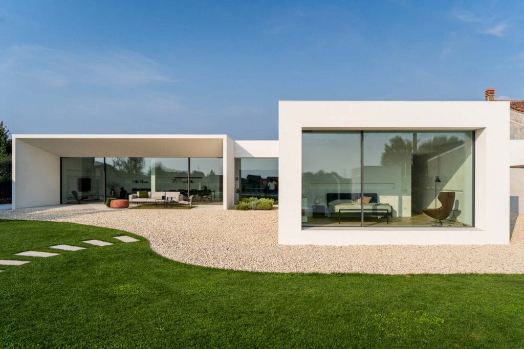 Immagine di una moderna villa con grandi finestre in vetro, situata in un'area verde con prato e ciottoli.