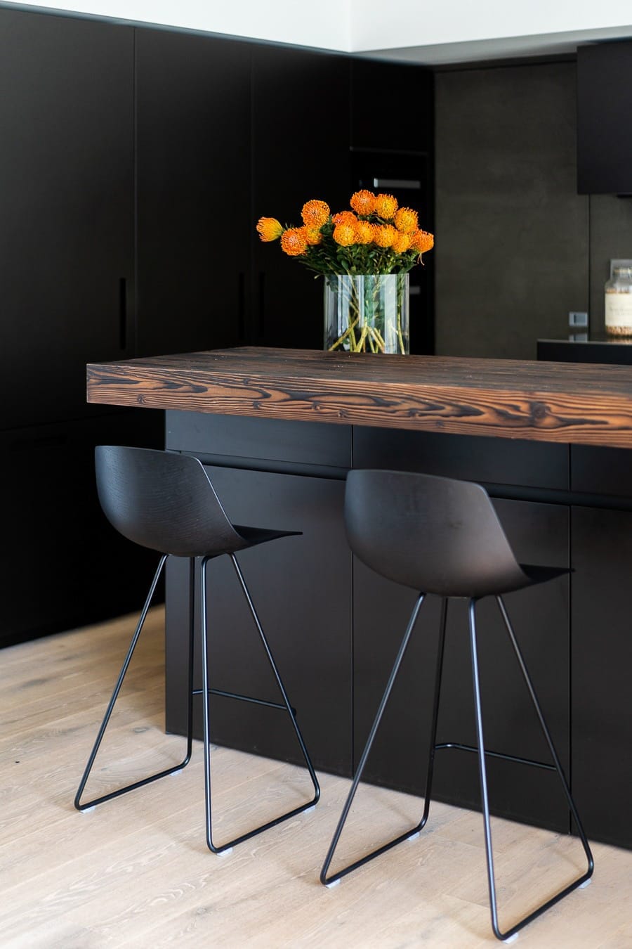 Dettaglio dell'isola cucina a Villa MD Home, Verona, con piano in legno massello, mobili neri minimalisti, sgabello di design e vaso di fiori arancioni. Design di lusso firmato Federico Cappellina & Partners Architects