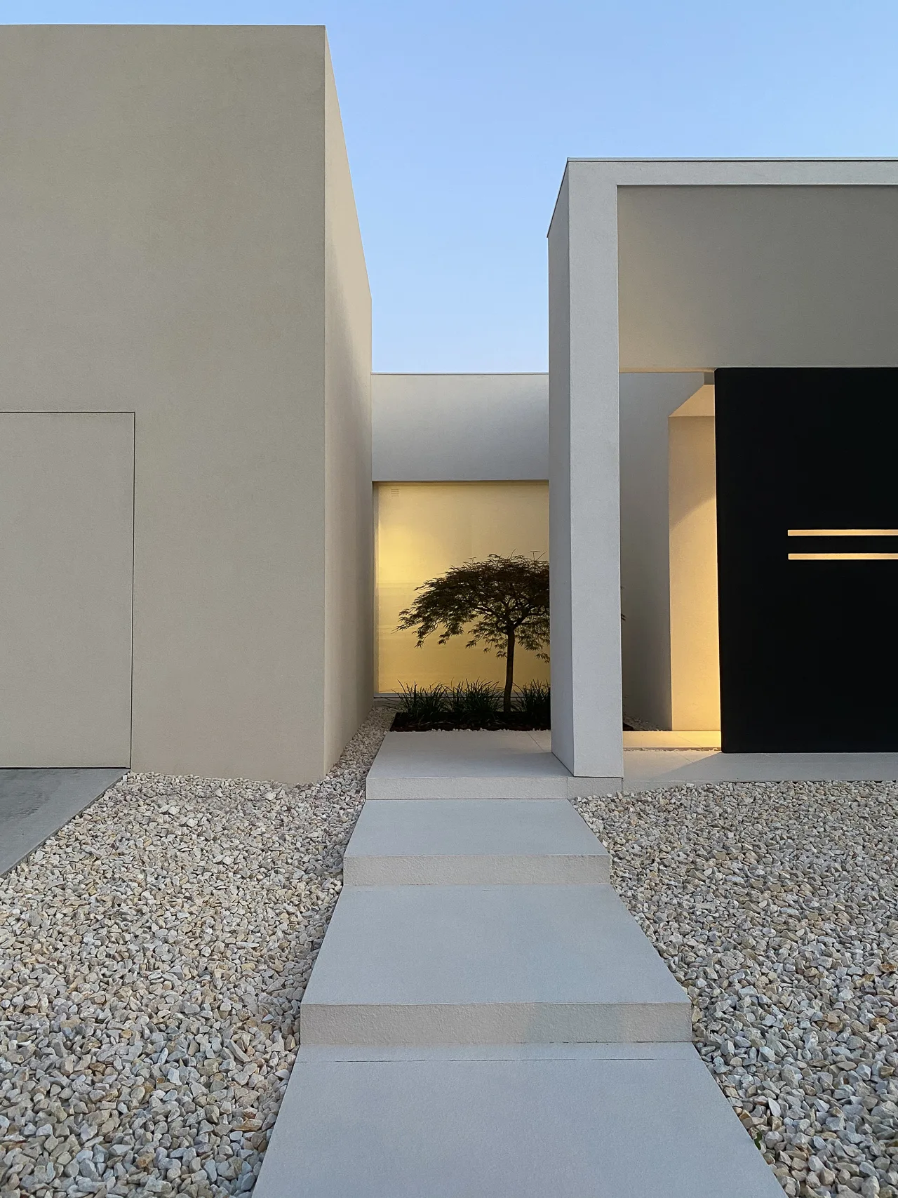 Architettura minimalista di Villa MD Home, Verona, con porta nera a filo muro, pareti bianche, tetto triangolare a sbalzo, ghiaia chiara, vegetazione ornamentale e albero giapponese. Design esclusivo di Federico Cappellina & Partners Architects