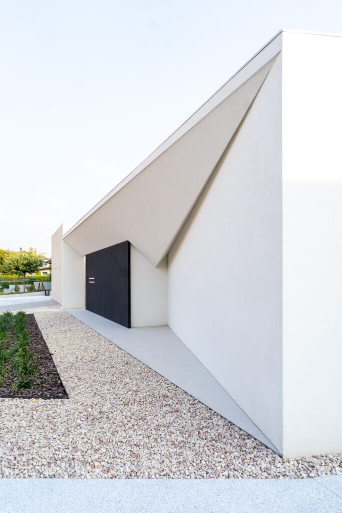 Architettura minimalista di Villa MD Home, Verona, con porta scura a filo muro, pareti bianche, tetto triangolare a sbalzo, ghiaia chiara e vegetazione. Design esclusivo di Federico Cappellina & Partners Architects