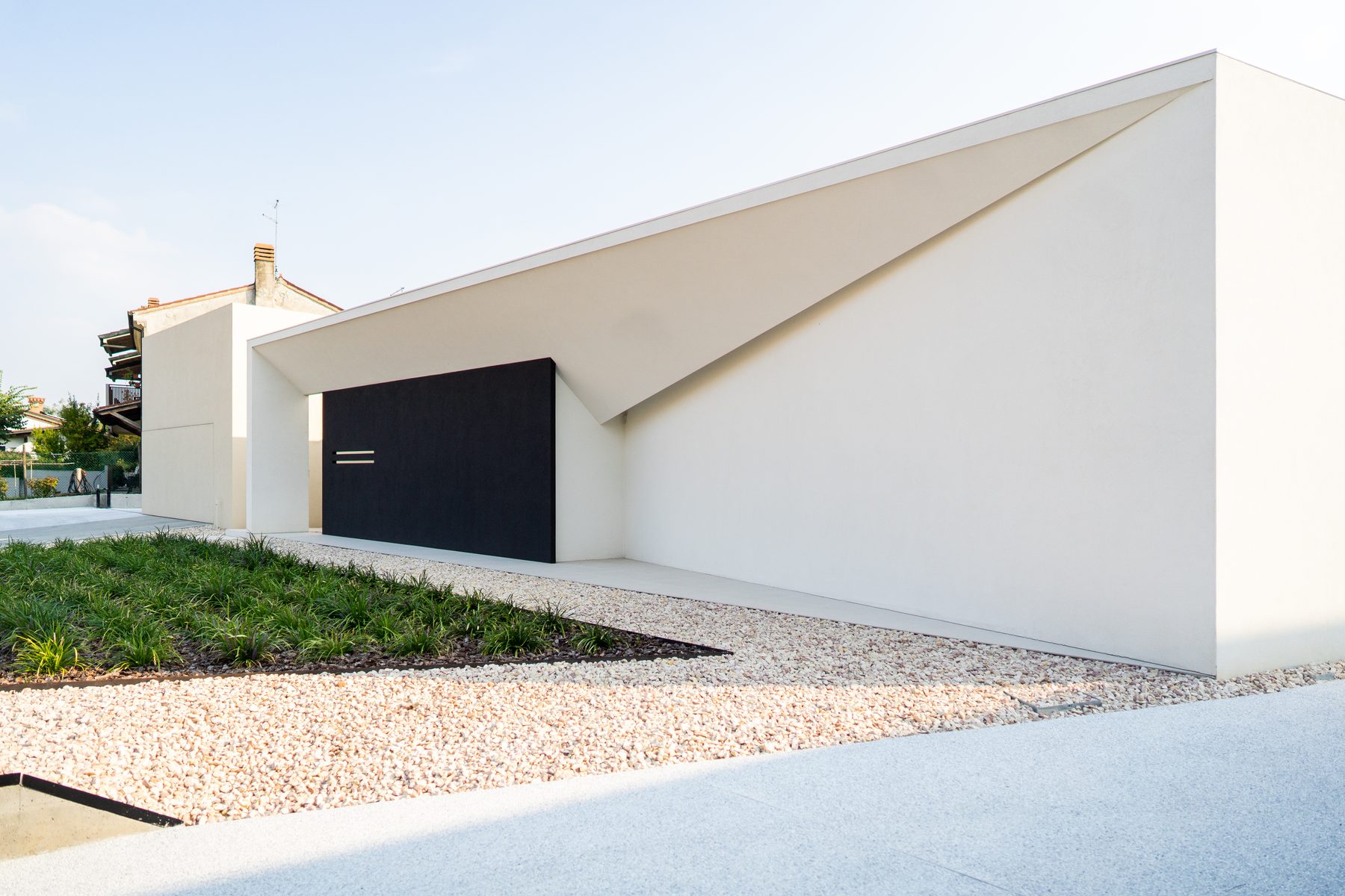 Fronte della villa verso la strada: parete bianca, ghiaia chiara e porta nera a filo muro